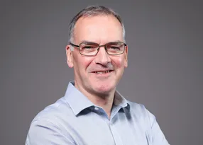 Neutral portrait of a person with short grey hair and glasses, wearing a light blue shirt, smiling slightly, against a plain grey backdrop.