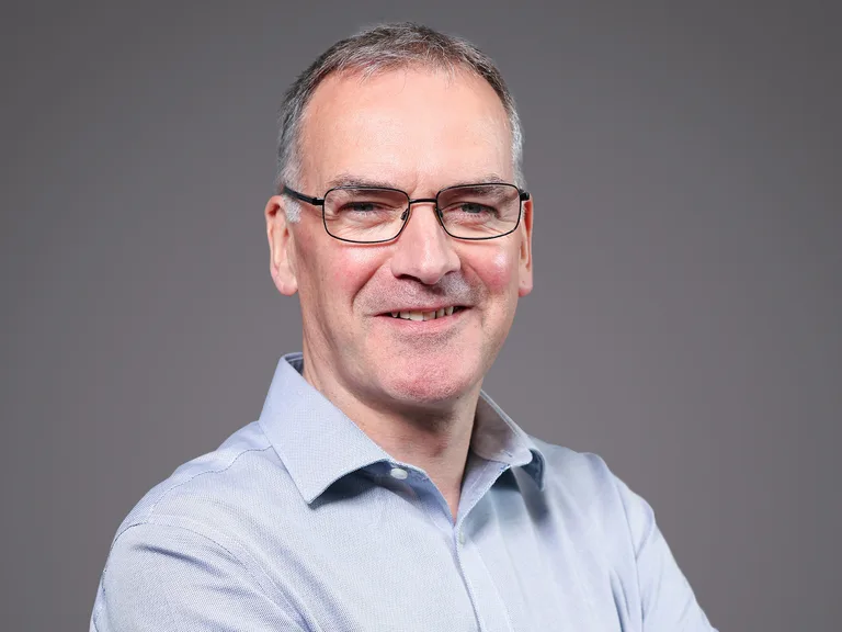 Neutral portrait of a person with short grey hair and glasses, wearing a light blue shirt, smiling slightly, against a plain grey backdrop.