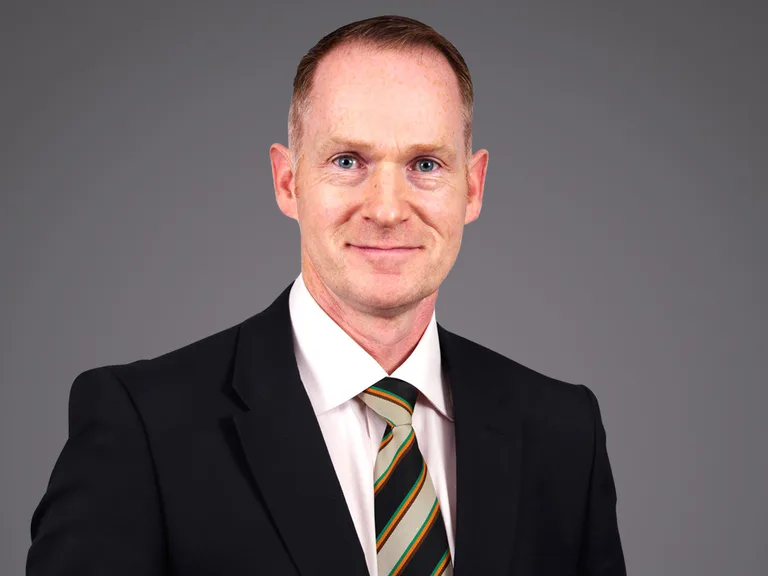 Primo piano di una persona in completo nero con camicia bianca e cravatta a righe verde, arancione e beige; sfondo grigio neutro.