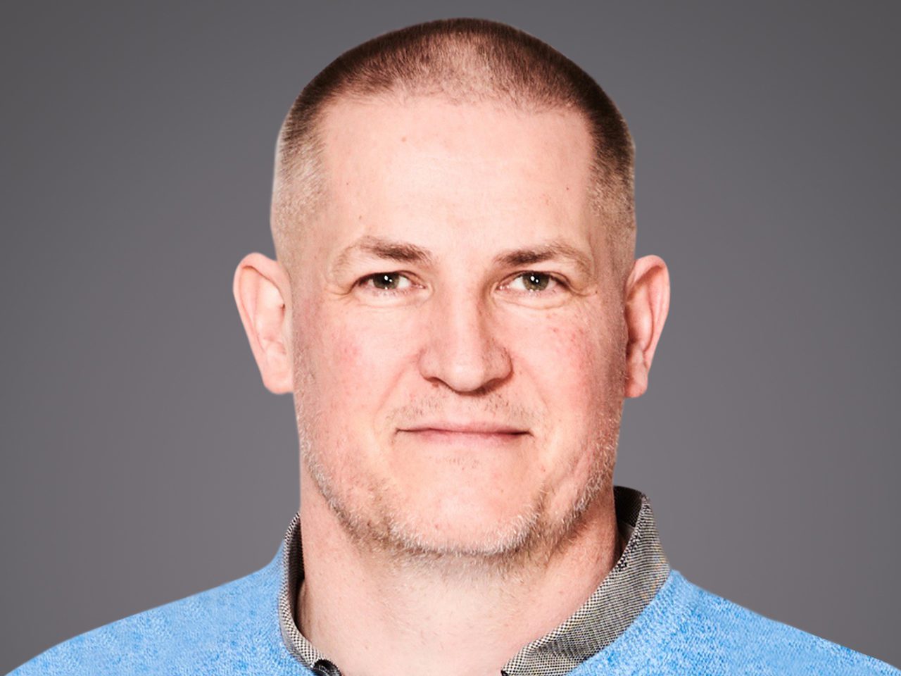 Head-and-shoulders portrait of a person with a close-cropped haircut and light stubble, wearing a light blue sweater over a collared shirt, against a grey background.