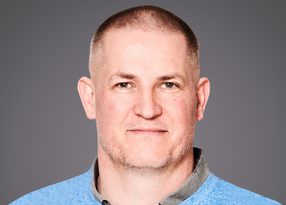 Head-and-shoulders portrait of a person with a close-cropped haircut and light stubble, wearing a light blue sweater over a collared shirt, against a grey background.
