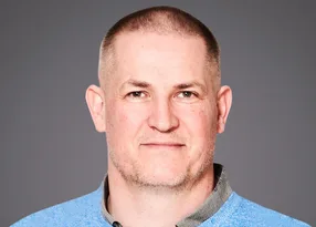 Head-and-shoulders portrait of a person with a close-cropped haircut and light stubble, wearing a light blue sweater over a collared shirt, against a grey background.