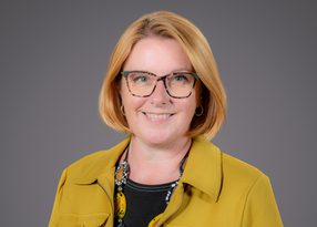 Portrait of a smiling person with short blonde hair and tortoiseshell glasses, wearing a mustard-yellow jacket over a black top and a beaded necklace.