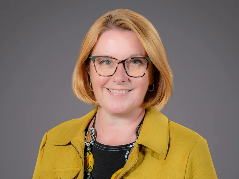 A smiling woman with red hair, wearing glasses, earrings, a black and yellow patterned scarf, and a yellow jacket against a grey background.