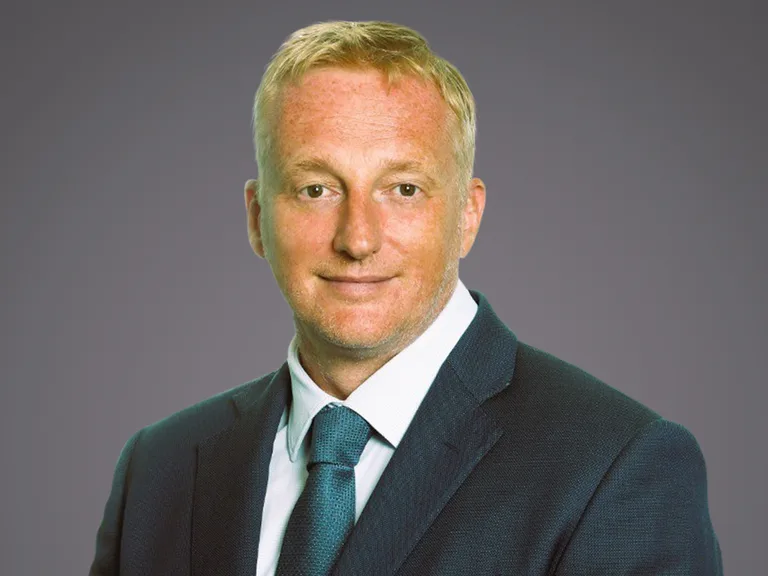 Head-and-shoulders portrait of a person with light blond hair, wearing a dark suit and teal tie, smiling softly against a plain grey background.