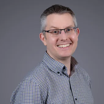Portrait of a smiling person with short grey hair and glasses, wearing a blue and white check shirt, against a smooth grey gradient background.