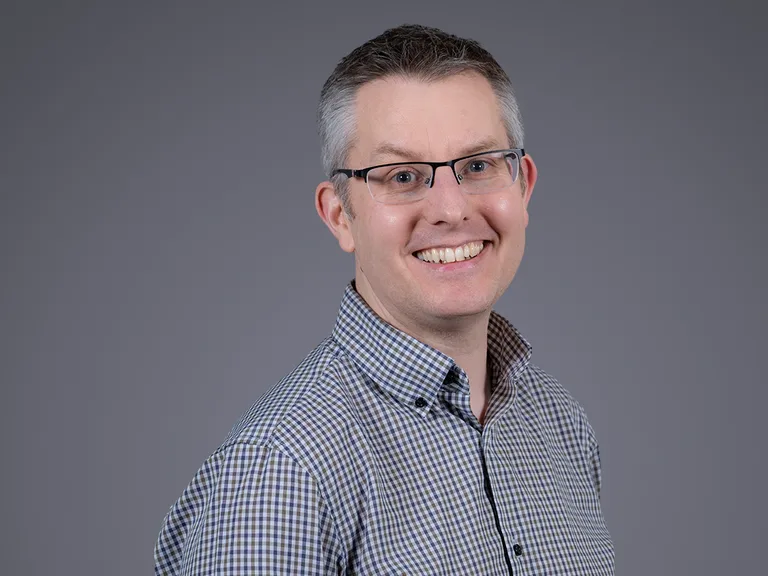 Portrait of a smiling person with short grey hair and glasses, wearing a blue and white check shirt, against a smooth grey gradient background.