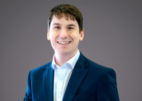 Portrait of a person with short dark hair, wearing a navy blue suit and white shirt, smiling against a neutral grey background.