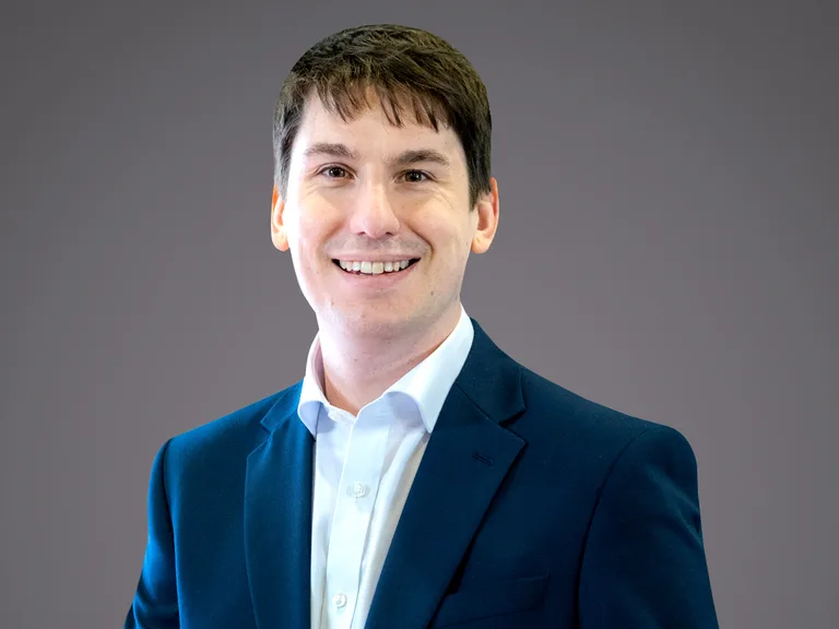 Portrait of a person with short dark hair, wearing a navy blue suit and white shirt, smiling against a neutral grey background.