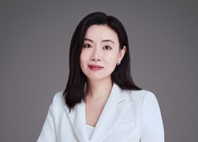 Portrait of a person with shoulder-length black hair, wearing a light blazer, facing the camera with a gentle smile against a grey studio background.