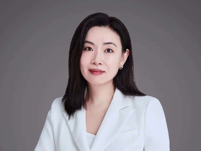 Portrait of a person with shoulder-length black hair, wearing a light blazer, facing the camera with a gentle smile against a grey studio background.
