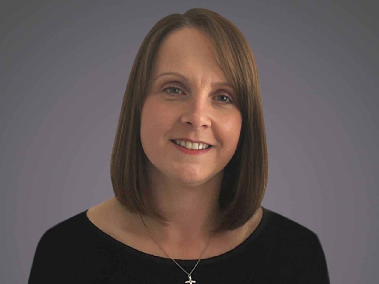 Smiling person with shoulder-length light brown hair, wearing a black top and a silver cross necklace, against a grey studio background.