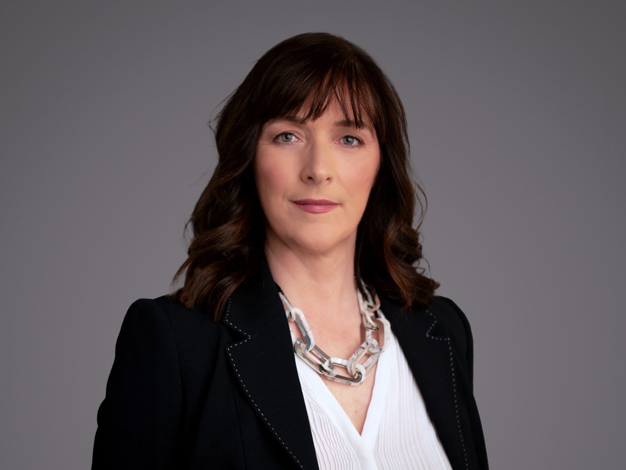 Portrait of a person with shoulder-length brown hair, wearing a black blazer, white blouse, and chunky silver necklace, against a gray backdrop.