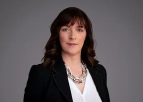 Portrait of a person with shoulder-length dark brown hair, wearing a black blazer over a white blouse and a chunky silver necklace, against a grey studio backdrop.