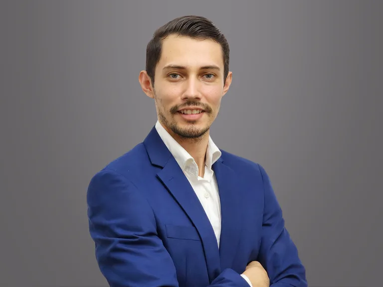 Une personne en veste bleue et chemise blanche, bras croisés, sourire léger, sur un fond gris dégradé.