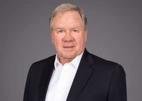 Head-and-shoulders portrait of a person with light hair, wearing a white shirt and dark blazer, with a neutral expression against a grey backdrop.
