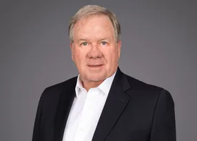 Head-and-shoulders portrait of a person with light hair, wearing a white shirt and dark blazer, with a neutral expression against a grey backdrop.