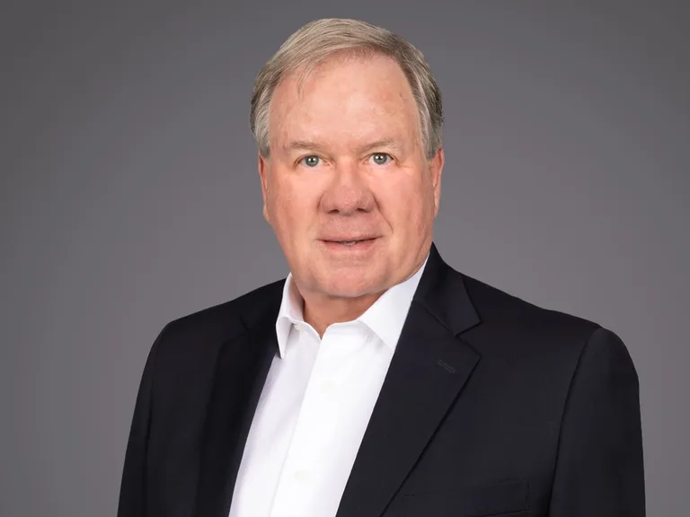 Head-and-shoulders portrait of a person with light hair, wearing a white shirt and dark blazer, with a neutral expression against a grey backdrop.