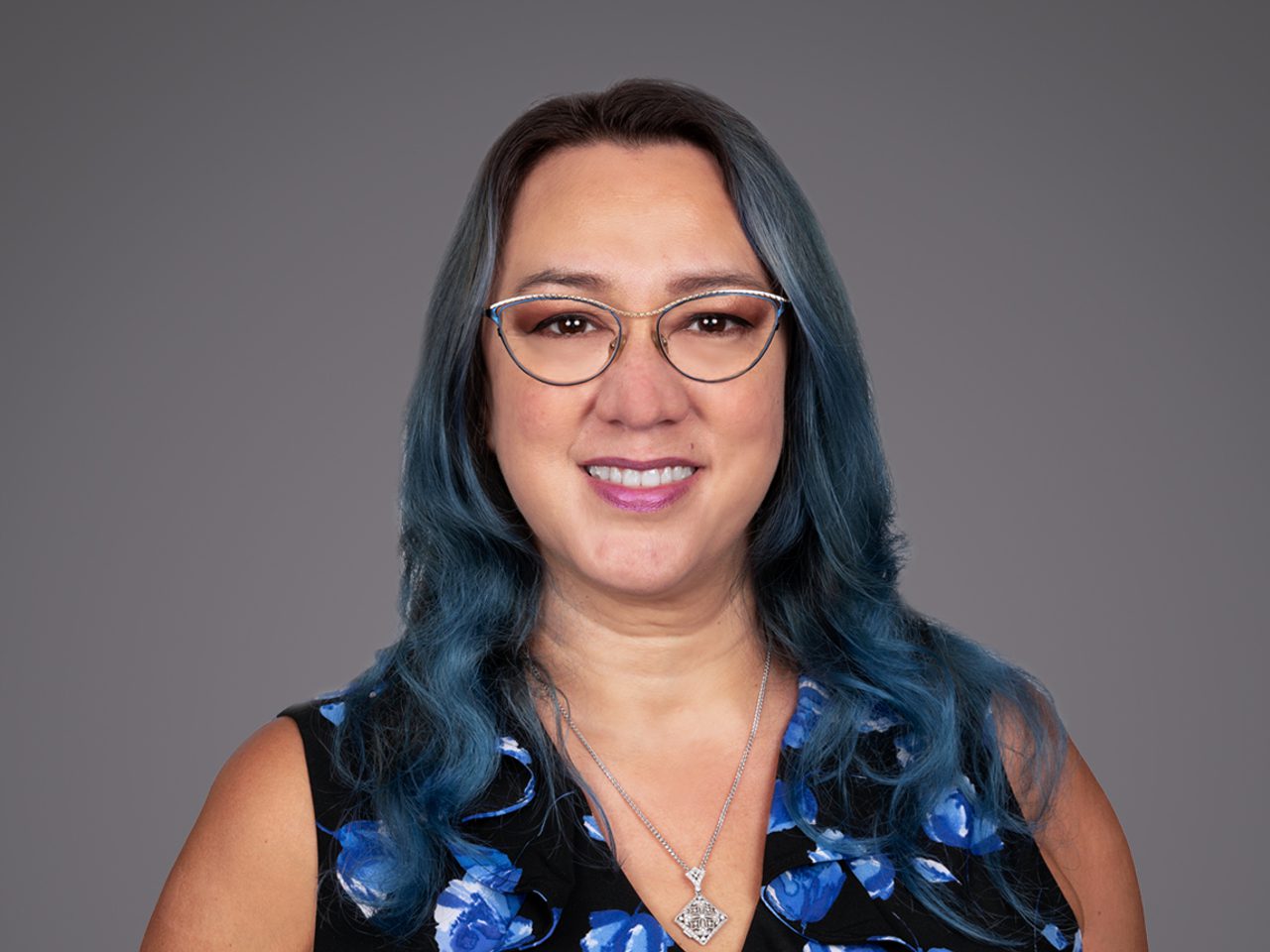 Portrait of a person with wavy blue-dyed hair, wearing round glasses, a black top with blue floral pattern, and a silver pendant, smiling against a grey backdrop.