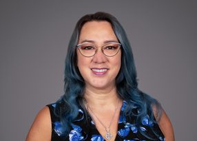 Portrait of a person with wavy blue-dyed hair, wearing round glasses, a black top with blue floral pattern, and a silver pendant, smiling against a grey backdrop.