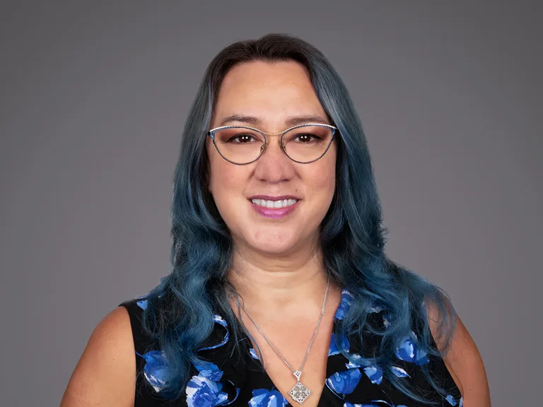 Portrait of a person with blue-green hair and glasses, wearing a blue floral top and a silver pendant, smiling against a gray studio backdrop.