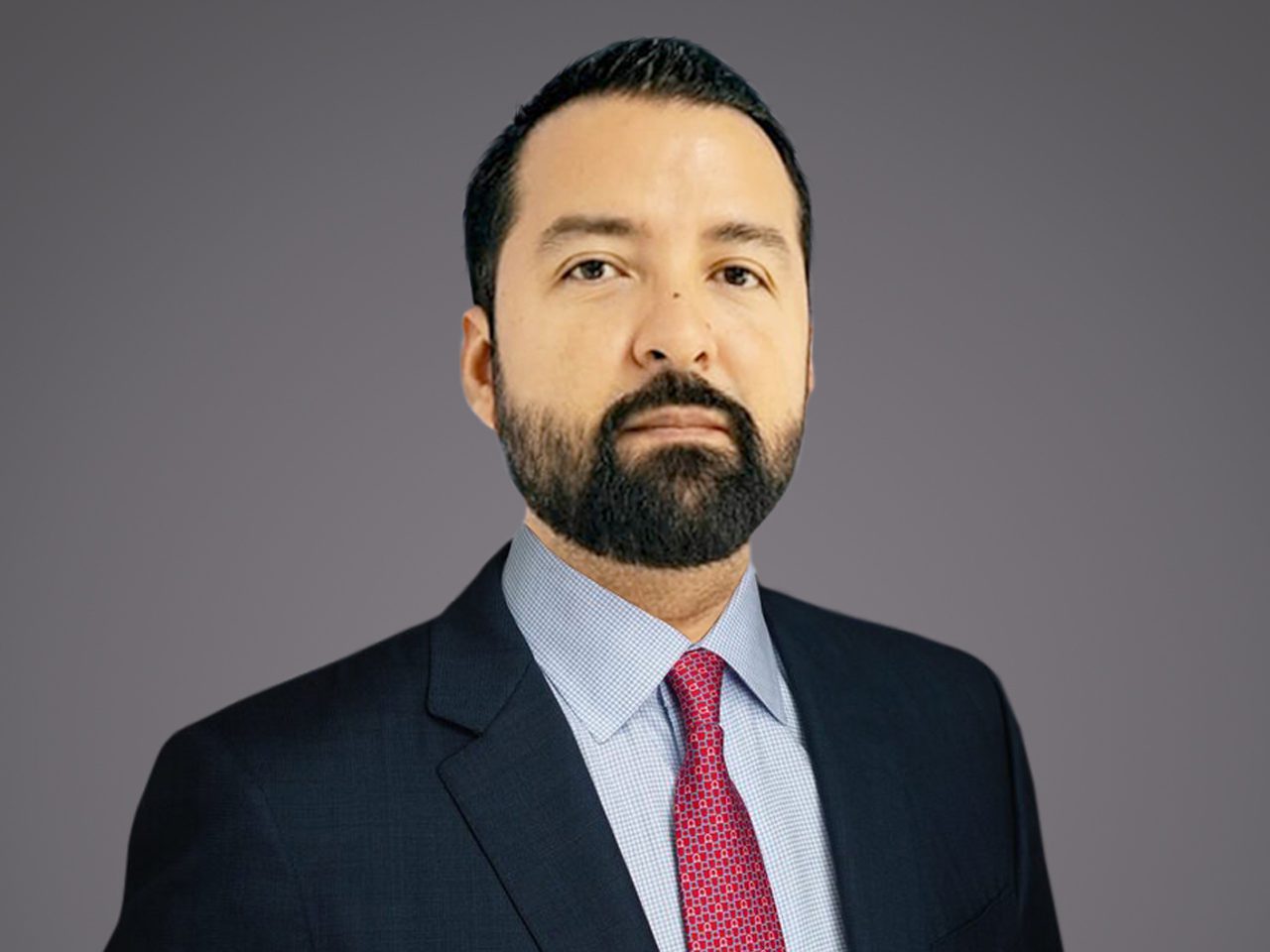 Persona con barba scura e capelli neri, in completo blu scuro, camicia azzurra e cravatta rossa, sfondo grigio uniforme.