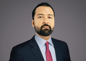 Personne adulte en costume sombre, chemise bleu clair et cravate rouge, barbe complète et regard droit, fond gris neutre.