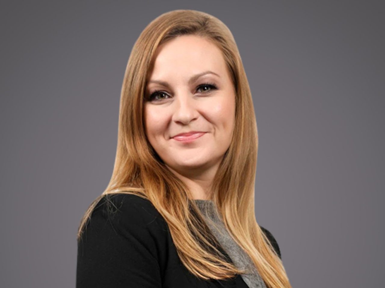 Portrait of a person with long blonde hair, wearing a black blazer over a grey top, smiling against a grey gradient background.
