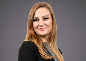 Portrait of a person with long blonde hair, wearing a black blazer over a grey top, smiling against a grey gradient background.
