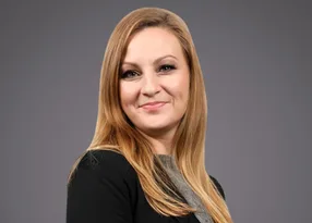 Portrait of a person with long blonde hair, wearing a black blazer over a grey top, smiling against a grey gradient background.