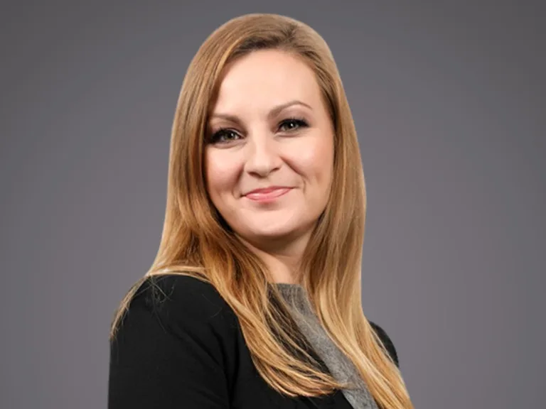 Portrait of a person with long blonde hair, wearing a black blazer over a grey top, smiling against a grey gradient background.
