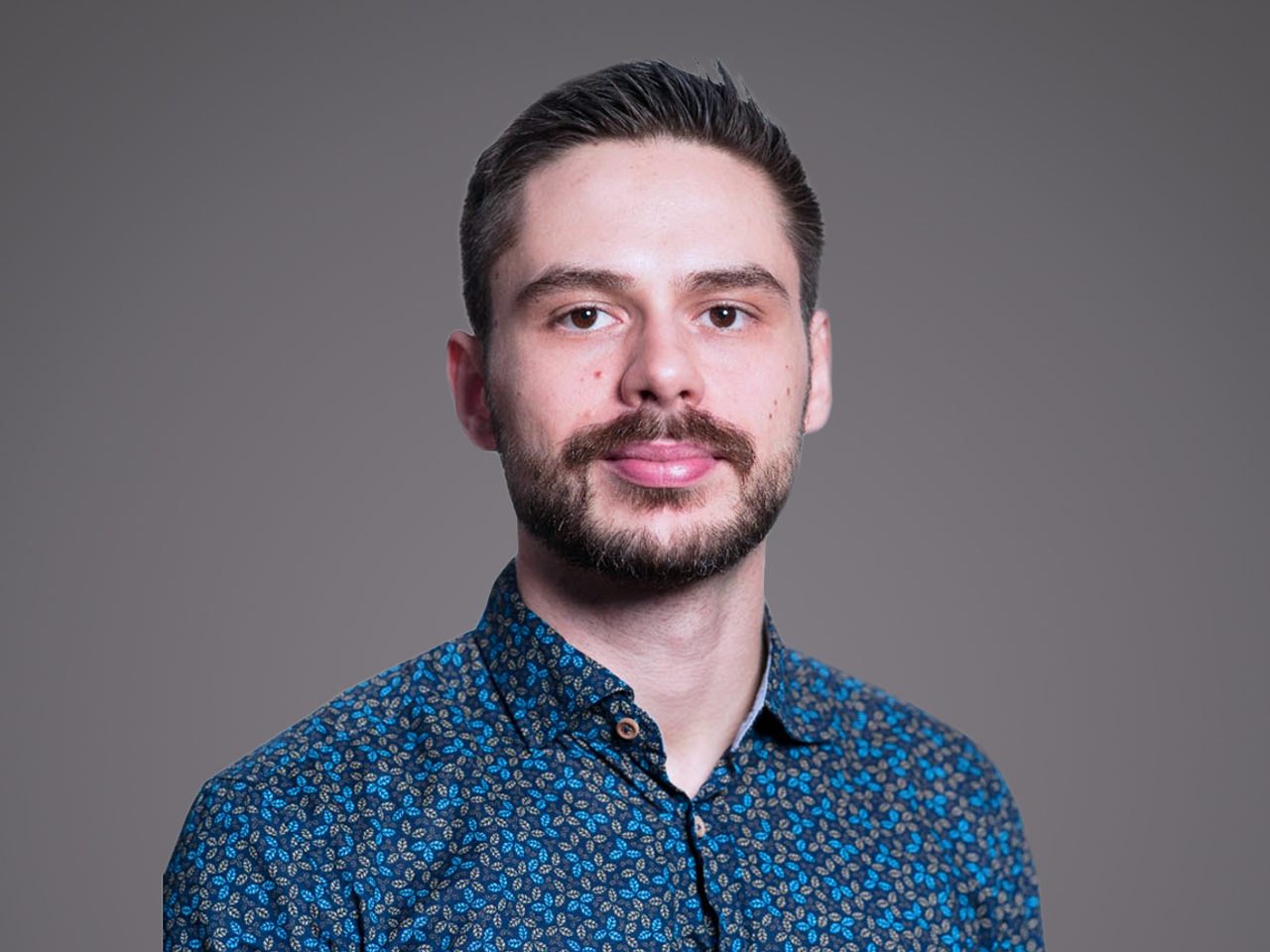 Portrait d'une personne avec barbe et cheveux courts, portant une chemise bleue à motif discret, fond gris uni.
