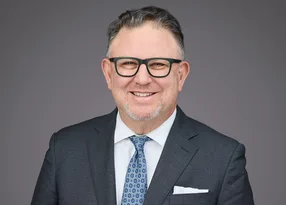 Portrait of a smiling person with short gray hair and dark-framed glasses, wearing a dark suit, white shirt, blue patterned tie, and a pocket square.