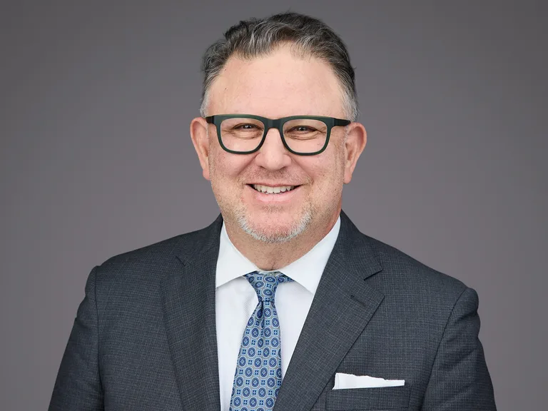 Porträt einer Person in dunklem Anzug, weißem Hemd und blau gemusterter Krawatte; Brille, Einstecktuch, grauer Hintergrund; lächelnd.