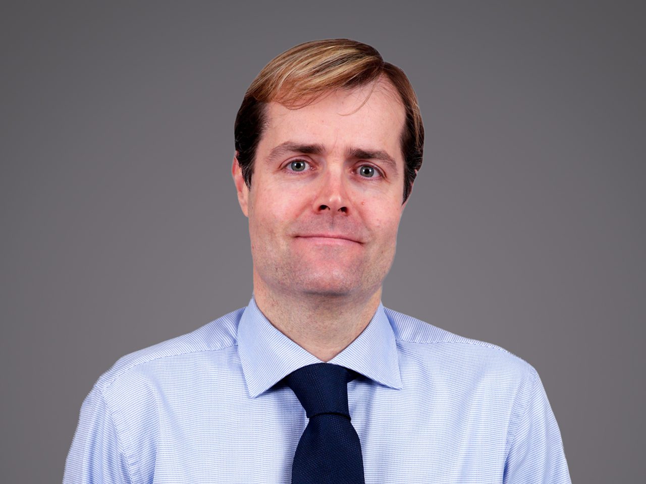 A person with light brown hair wearing a pale blue shirt and navy tie, against a grey background, facing the camera with a gentle smile.