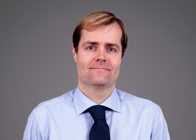 A person with light brown hair wearing a pale blue shirt and navy tie, against a grey background, facing the camera with a gentle smile.