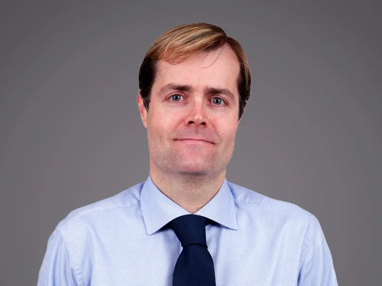 A person with light brown hair wearing a pale blue shirt and navy tie, against a grey background, facing the camera with a gentle smile.