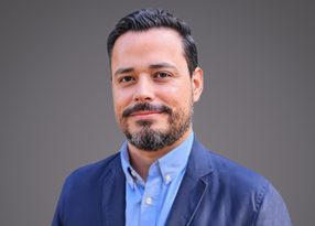 Personne souriante portant une veste bleue et une chemise bleu clair, cheveux courts et barbe, sur fond gris dégradé.