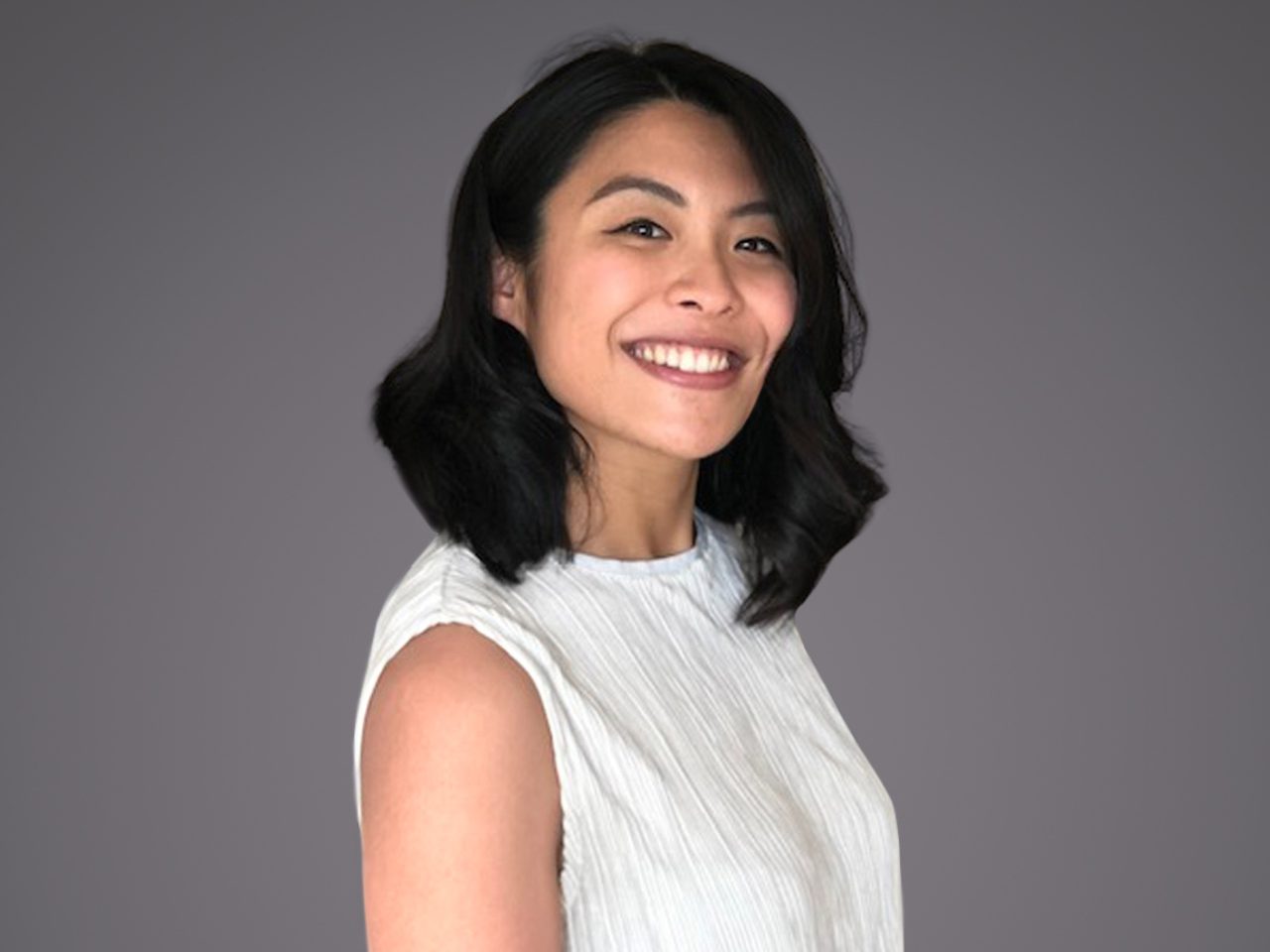 Person with shoulder-length dark hair, wearing a white sleeveless top, smiling at the camera against a neutral grey background.