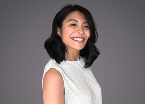 Personne souriante aux cheveux noirs mi-longs, vêtue d'un haut blanc sans manches, sur fond gris.