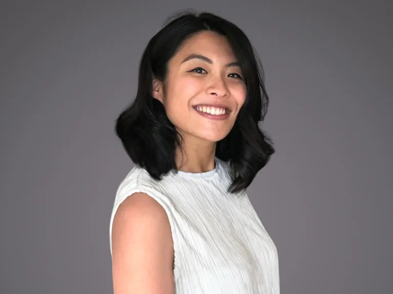 Person with shoulder-length dark hair, wearing a white sleeveless top, smiling at the camera against a neutral grey background.