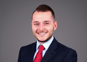 Professional portrait of a person with short hair, wearing a navy suit, light blue shirt and red tie, smiling against a grey studio backdrop.