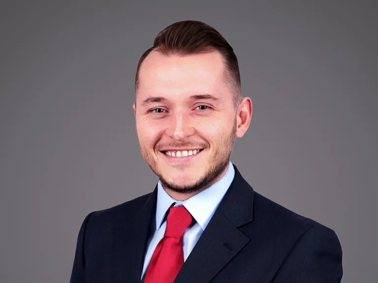 Professional portrait of a person with short hair, wearing a navy suit, light blue shirt and red tie, smiling against a grey studio backdrop.