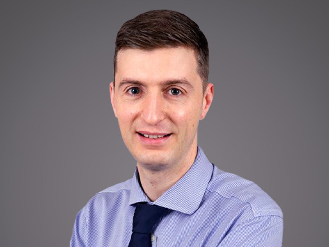 Person with short dark hair, wearing a light blue shirt and dark tie, smiling against a grey studio background.