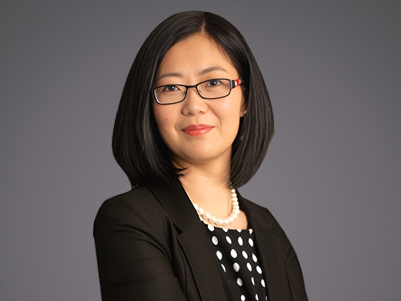 Portrait of a person with shoulder-length black hair and glasses, wearing a black blazer, a polka-dot top, and a pearl necklace against a gray backdrop.