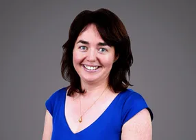 Smiling person with shoulder-length dark hair, wearing a cobalt-blue V-neck top and a pendant necklace, against a grey gradient backdrop.