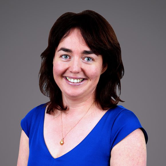 Smiling person with shoulder-length dark hair, wearing a cobalt-blue V-neck top and a pendant necklace, against a grey gradient backdrop.