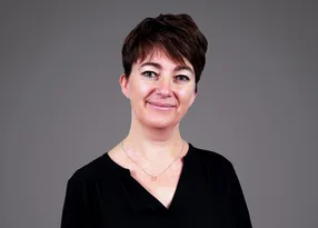 Person with short dark hair, wearing a black top and a silver necklace, smiles against a grey backdrop.
