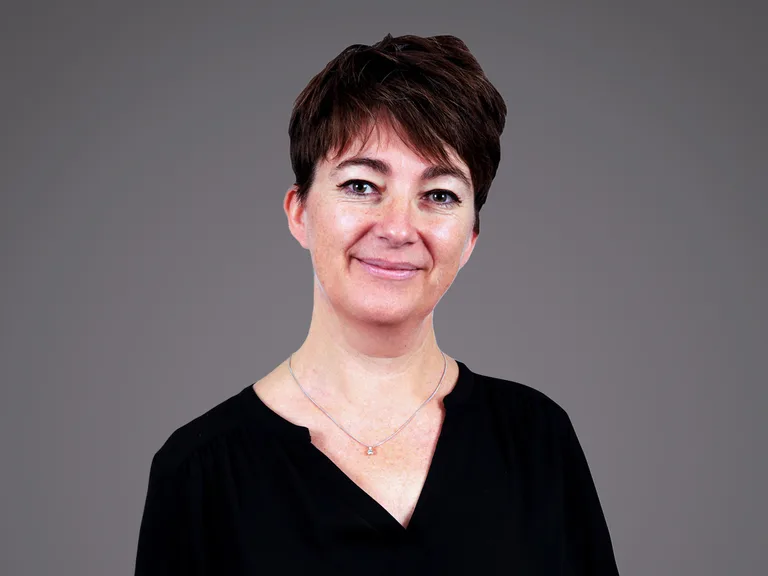 Portrait of a person with short dark hair, wearing a black top and a silver necklace, smiling softly against a neutral gray backdrop.