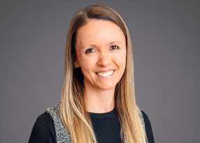 Head-and-shoulders portrait of a person with long light brown hair, smiling at the camera; wearing a black top with a textured grey vest, against a grey backdrop.